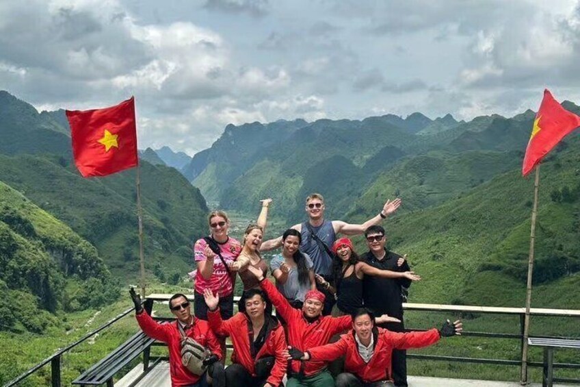 A group of people on a mountain ridge with flags, surrounded by green hills. 1