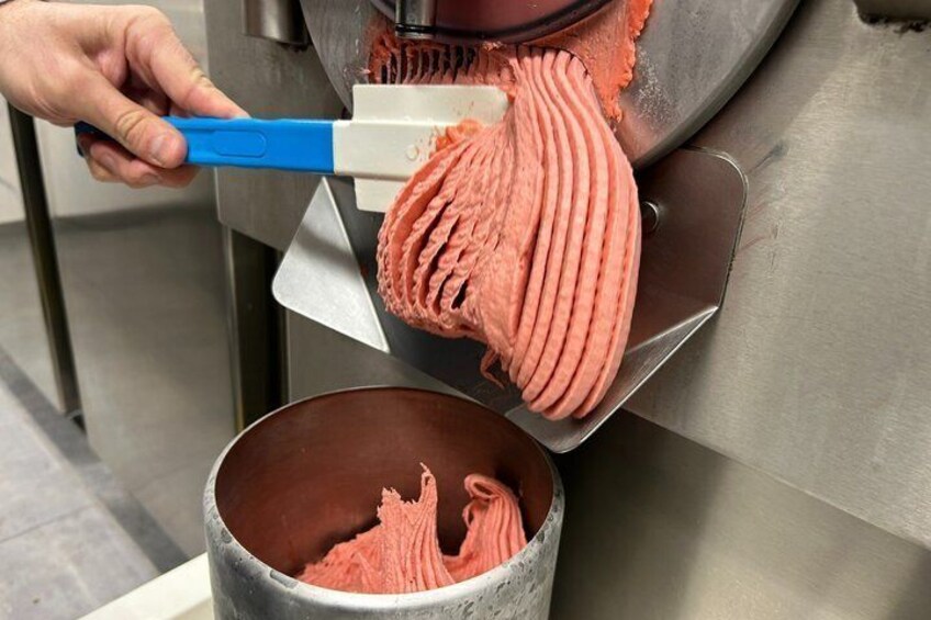 A person using a blue-handled tool to scrape ice cream from a metal machine into a metal container. 2