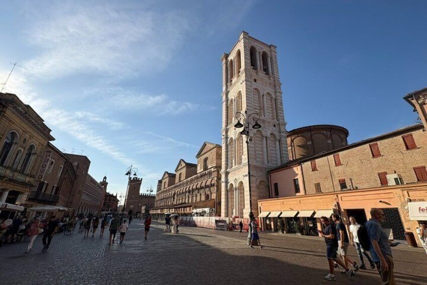 A cobblestone square with a tall bell tower, historic buildings, and people walking. 3