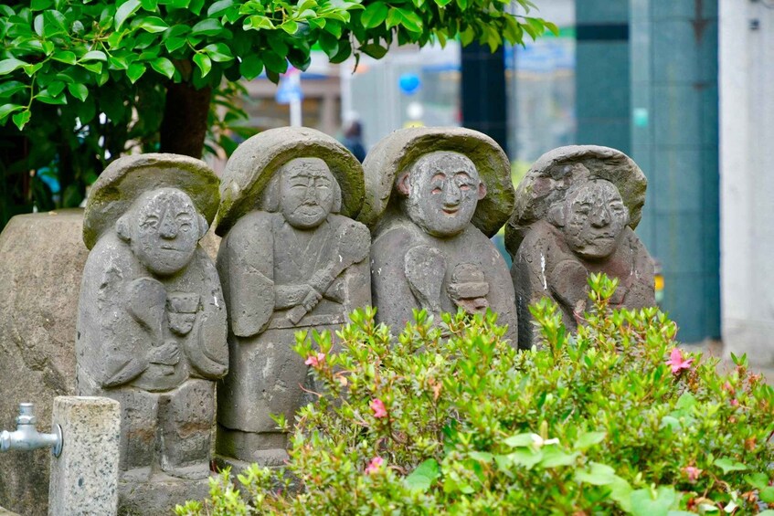 Stone statues of figures with hats and one holding a cup, set against a backdrop of greenery and a building. 5