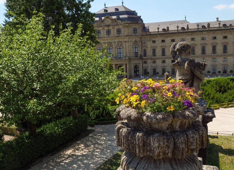 A stone statue of a figure with a flower, a garden with colorful flowers, and a large classical building in the background. 2