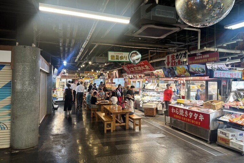 An indoor market with wooden tables and benches, food stalls, and people shopping. 3