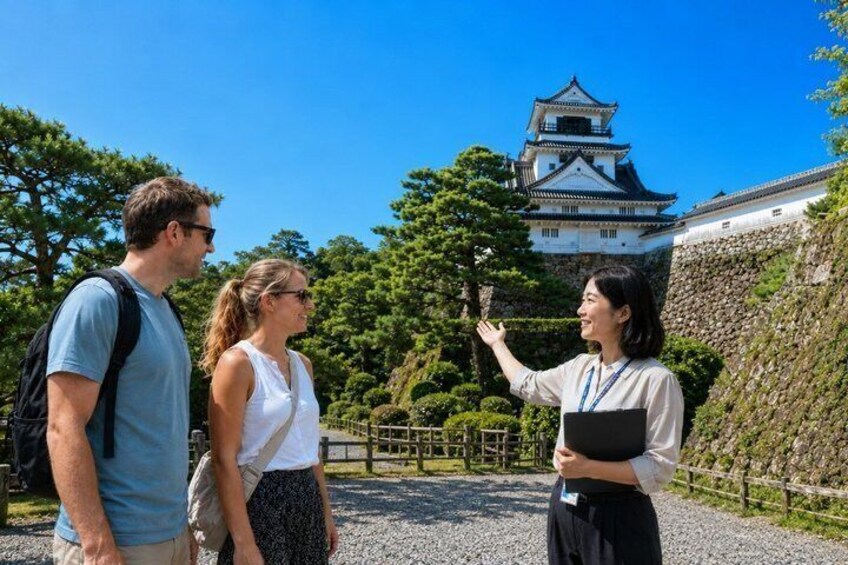 Three people standing in front of a castle with a traditional Japanese architectural style, a stone wall, and a gravel path. 1