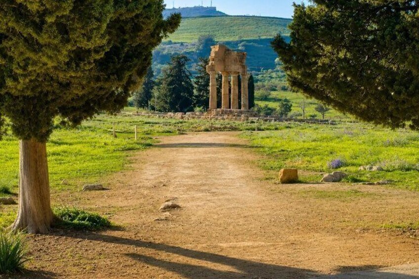 A dirt path leading to ancient ruins with columns, surrounded by trees and a hilly landscape. 5