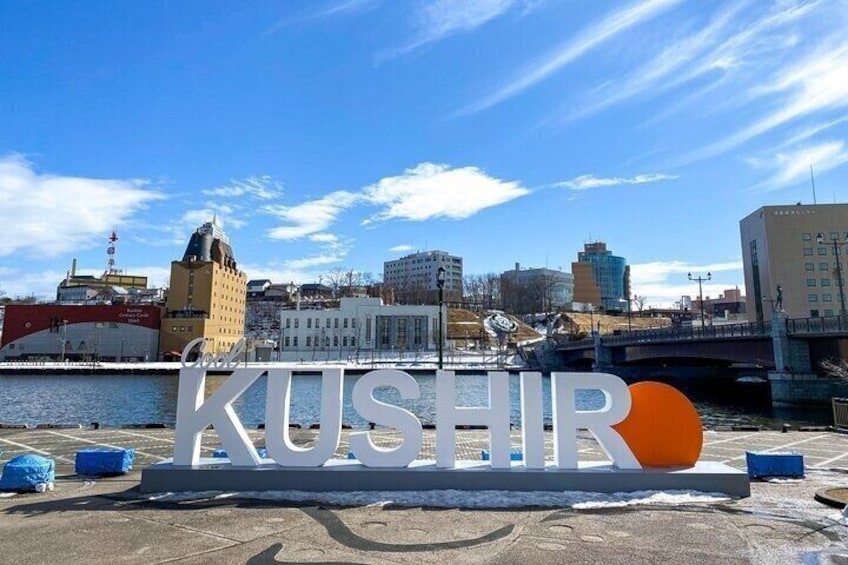 A large sign with the word 'KUSHIR' and a red circle, set against a backdrop of buildings and a river. 1