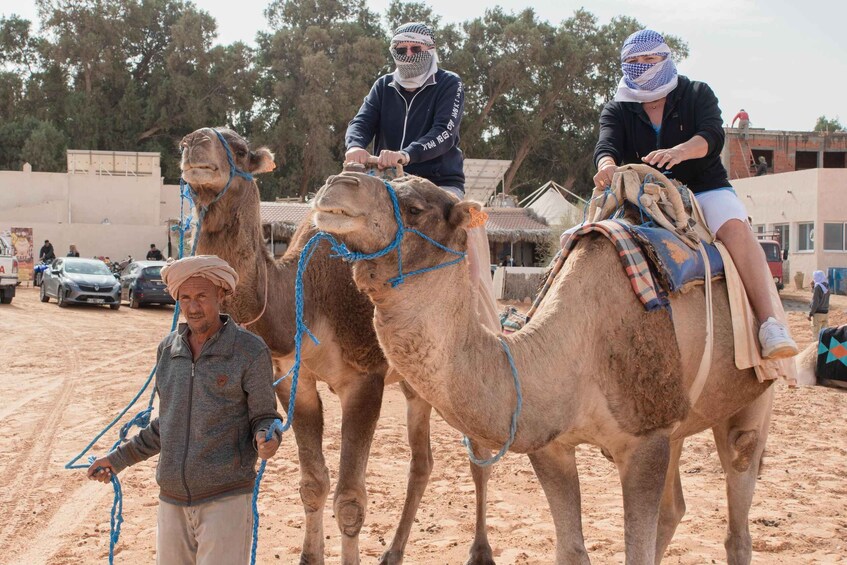 Three camels with riders, a sandy ground, and a few parked cars in the background. 3