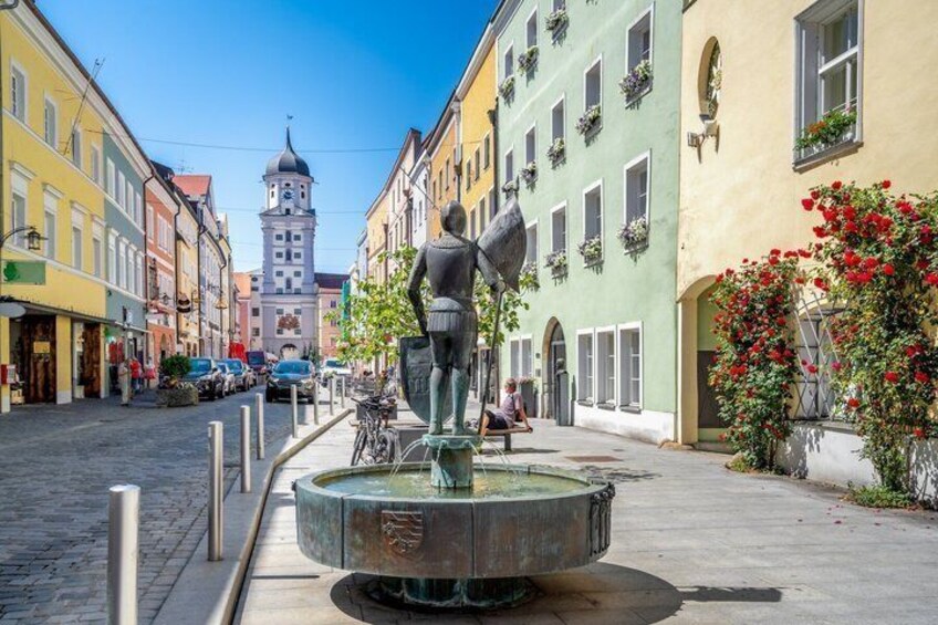 A cobblestone street with colorful buildings, a statue, and a tower in the background. 1