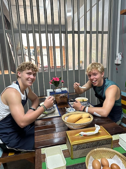 Two people sitting at a table with food and drink, a basket of bread, and a tissue box. 3