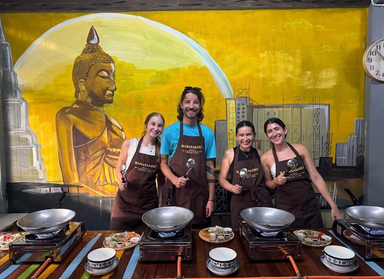 Four people wearing aprons with 'MAHANAKORN' on them, standing in front of a mural with a Buddha figure and a cityscape. 5