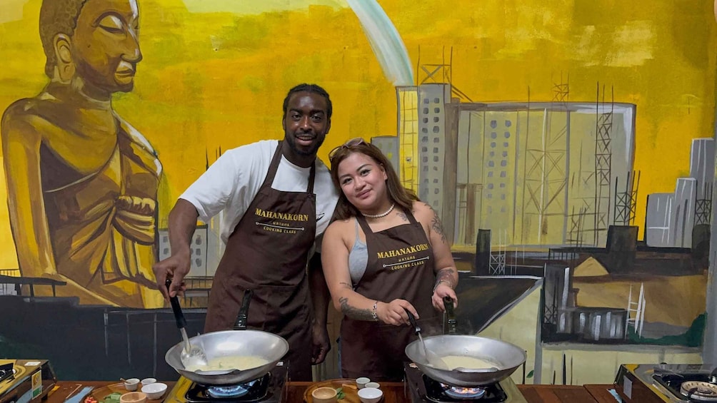 Two people in aprons cooking in a kitchen with a Buddha mural in the background. 4