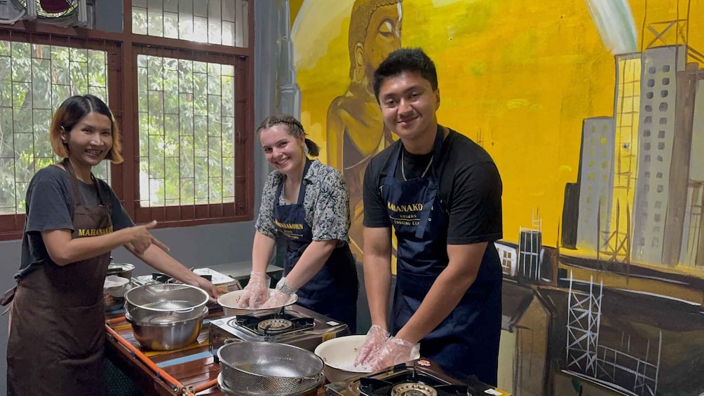 Three people in a kitchen, wearing aprons, with cooking utensils and pots on the counter. 2
