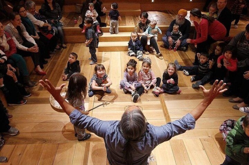 A group of children seated in a circle on a wooden floor, with an adult standing in front of them. 5