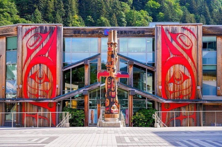 A totem pole in front of a building with large red designs. 1