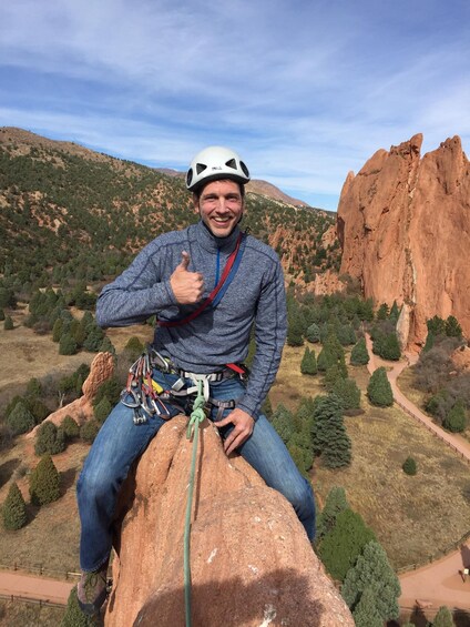 A person climbing a rock with a helmet and harness, surrounded by a mountainous landscape with trees and a clear sky. 3