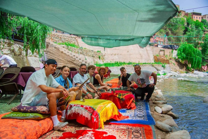 A group of people sitting on colorful rugs by a river, under a canopy. 2