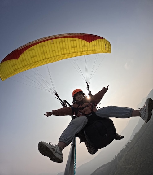 A person paragliding with a yellow and red parachute, wearing a helmet and harness, against a mountainous backdrop. 3