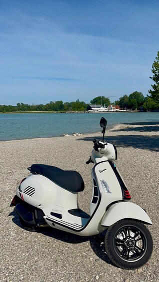 A white Vespa scooter parked on a pebbled beach with a lake and trees in the background. 2