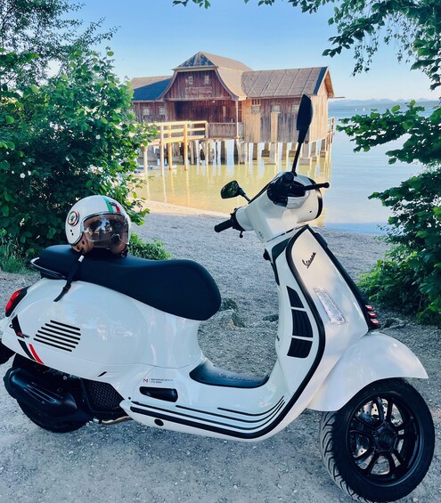 A white scooter with a helmet on the seat, parked on a gravel path with a wooden structure and a body of water in the background. 4