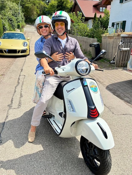 A white Vespa scooter parked on a paved surface with a yellow car in the background. 1