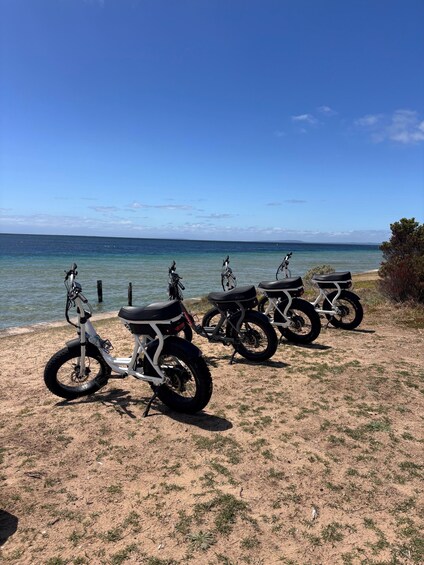 Four electric scooters parked on a sandy beach with the ocean in the background. 4