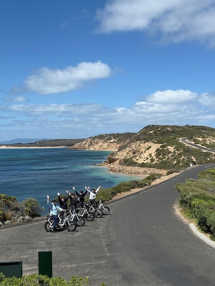 A coastal road with a group of cyclists on the side, a clear blue sky, and a hilly landscape in the background. 5