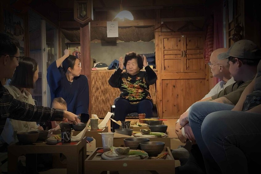 A group of people around a table with various dishes and utensils, in a room with wooden cabinets and a hanging light fixture. 1