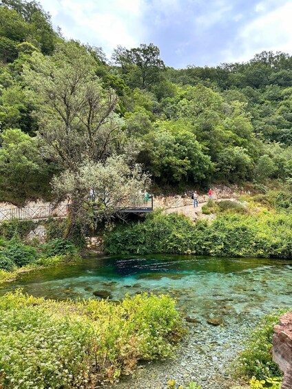A clear stream with people walking on a bridge and a forested hillside. 3