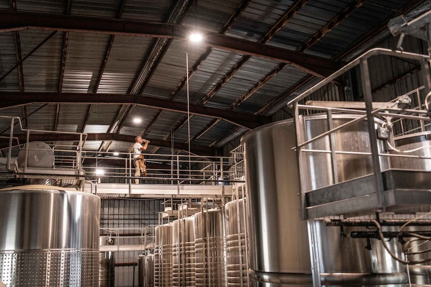 A person on a platform in a large industrial facility with multiple stainless steel tanks and metal structures. 3