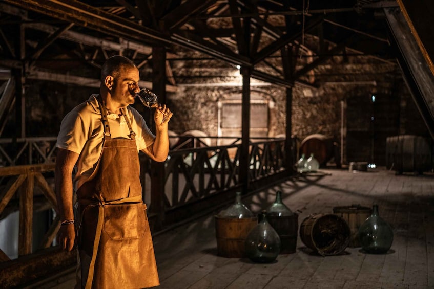 A person in an apron holding a glass, standing in a rustic interior with wooden beams and barrels. 5