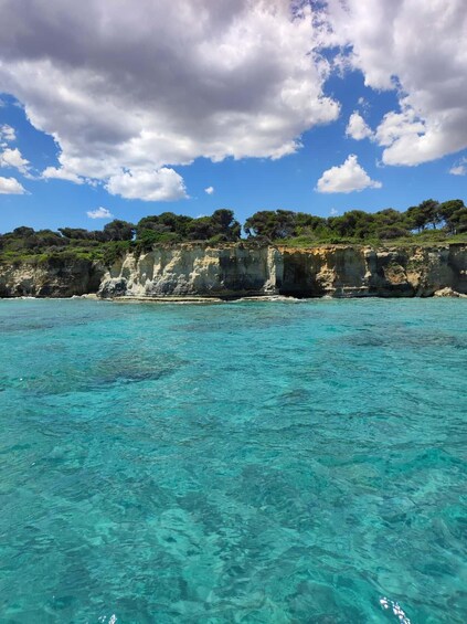 A coastal landscape with clear turquoise waters, rocky cliffs, and dense green vegetation under a partly cloudy sky. 3
