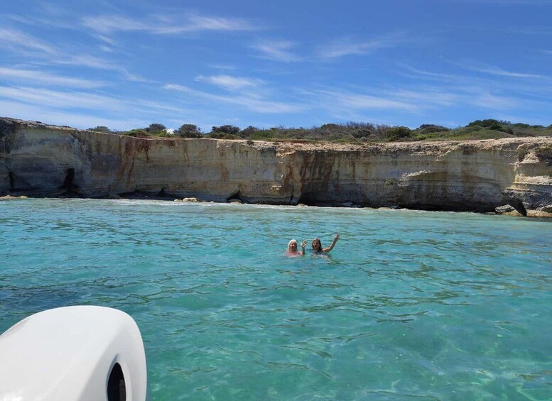 A clear blue sea with two people swimming, a cliff, and a boat. 2