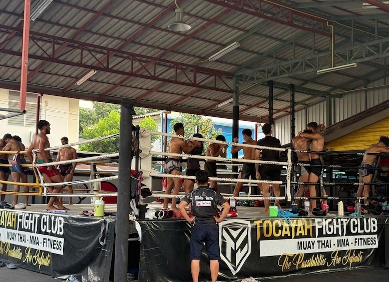 A boxing ring with fighters and trainers, surrounded by a covered area with banners for Tocaya Fight Club. 1