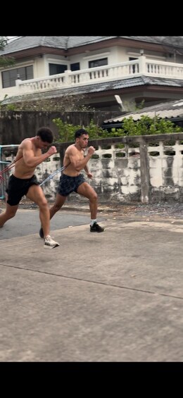 Two individuals running on a concrete path with a residential building and a white fence in the background. 3