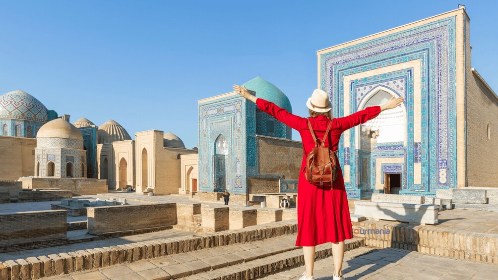 A person in a red dress and hat standing with arms outstretched in front of a building with blue and white tile patterns and domed structures. 3