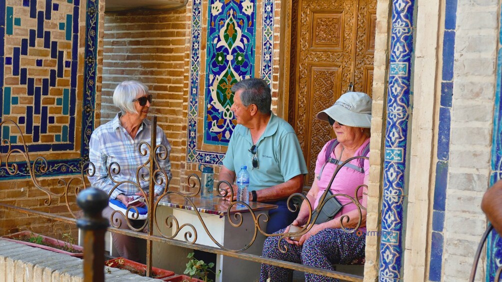 Three people sitting on a bench outside a building with intricate tile work. 5