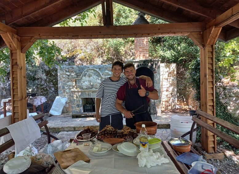 Two people standing in front of a stone fireplace under a wooden pavilion with a table full of food items. 3