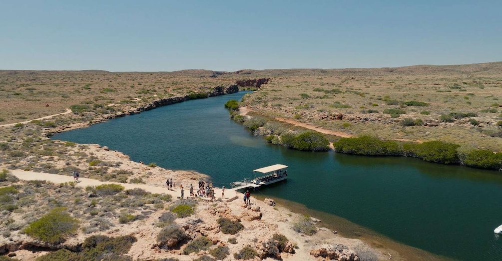 A river winding through a desert landscape with sparse vegetation, a boat docked at the riverbank, and people walking along the shore. 3