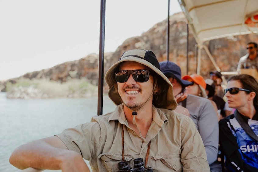 A person in a beige shirt and hat steering a boat, with rocky cliffs and calm water in the background. 4