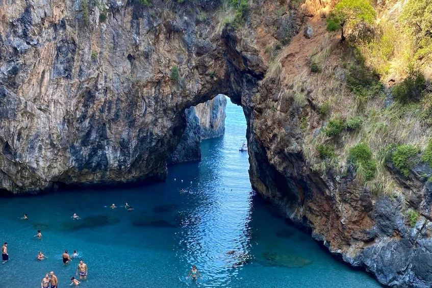 A natural rock arch over a clear blue water body with people swimming and floating. 2