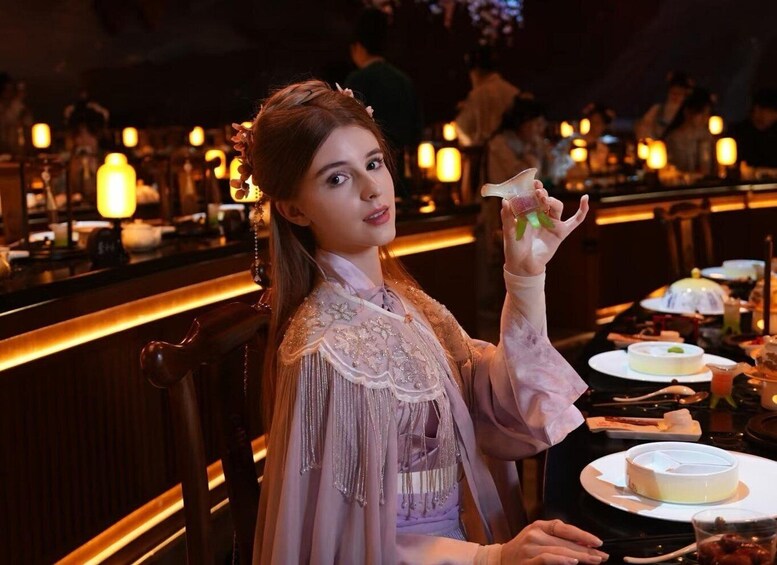 A person in traditional attire holding a flower, seated at a table with a dessert and a drink. 1