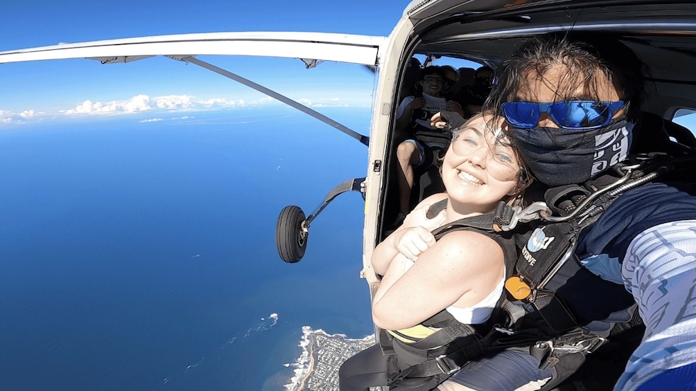 A person in a parachute harness inside a small aircraft, with a view of the ocean and coastline below. 4