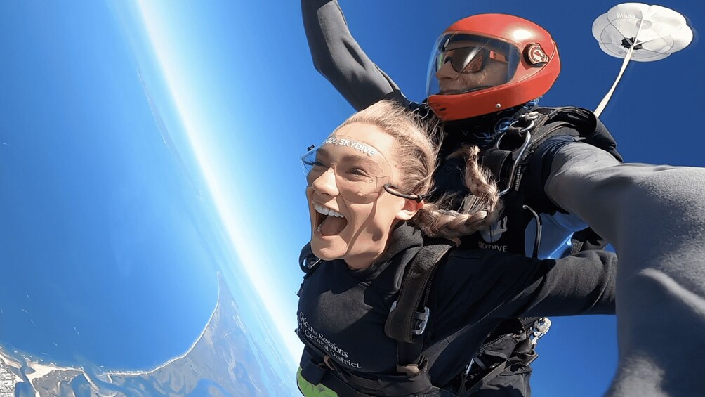 A person skydiving with a parachute, another person taking a selfie, and a clear blue sky. 1