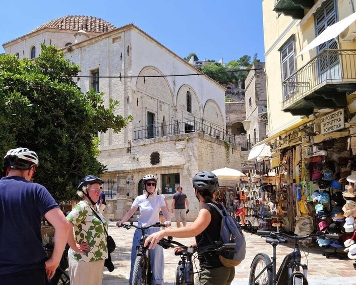 A group of cyclists on a street with shops and a historic building in the background. 1