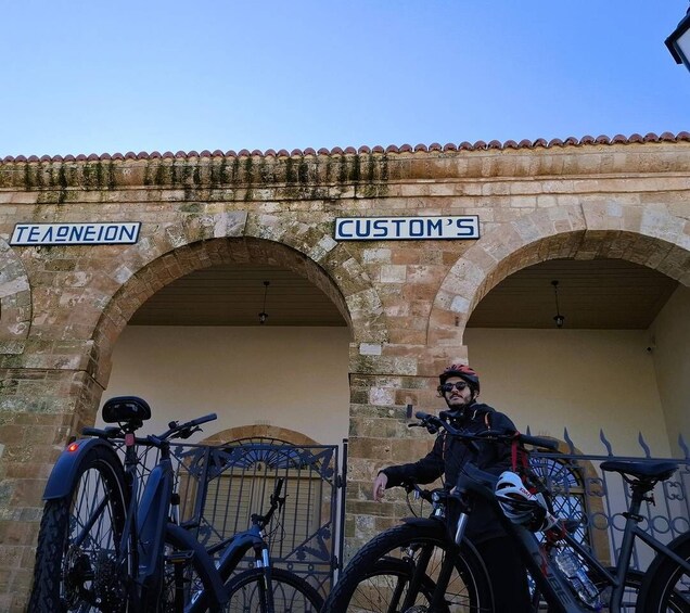 A person standing next to a bicycle in front of an old stone building with arches, labeled 'TEA NEITON' and 'CUSTOM'S.' 5