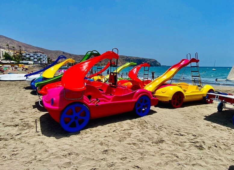 Colorful children's carousels on a sandy beach with a clear blue sky and a mountainous backdrop. 3