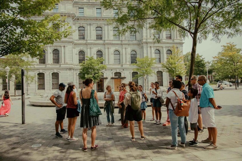 A group of people standing in a plaza in front of a large, historic building with arched windows and columns. 5