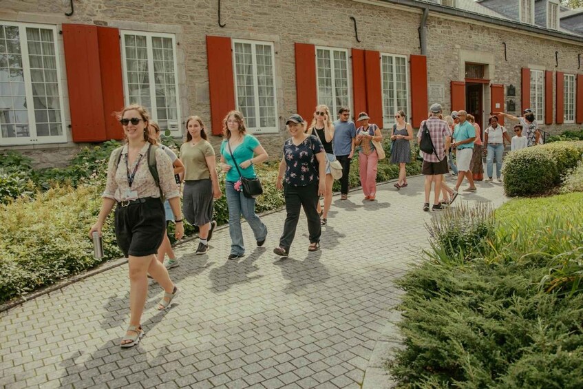 A group of people walking on a paved pathway in front of a building with red shutters and stone walls. 3