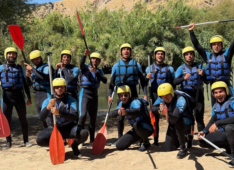 A group of people in blue life jackets and yellow helmets, some holding paddles, standing and kneeling by a river. 3