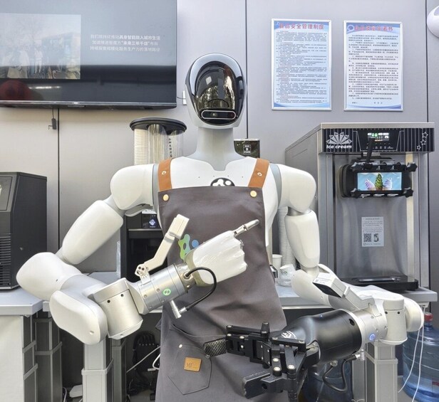 A mannequin with robotic arms and a helmet, wearing an apron, in a setting with a freezer and informational posters. 1