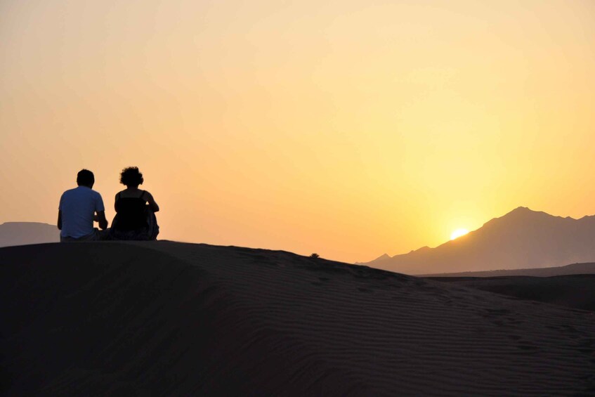 Two people sitting on a sand dune with a sunset in the background. 1